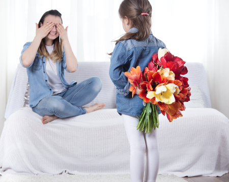 Happy mother's Day. Little sweet daughter with a large bouquet of tulips congratulates her mother. In the interior of the living room, the concept of a happy family lifeの写真素材
