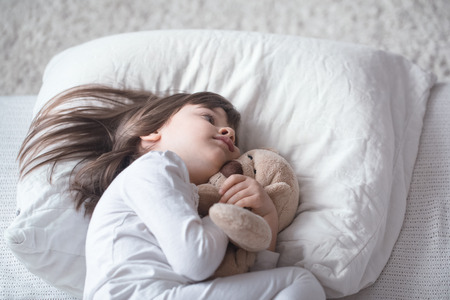 Cute little girl sleeps sweetly in a white cozy bed with a soft bear toy, the concept of children's rest and sleepの写真素材