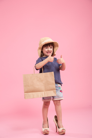 Funny little girl cute girl in summer hat with shopping bag and in the shoes of a mother, the concept of children's fashion, Studio shot isolated on pink backgroundの写真素材