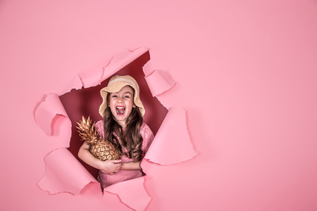 Funny little girl in a beach hat and with pineapple gold color, on a colored pink background, peeking out from the hole in the background, Studio shooting, space for textの写真素材