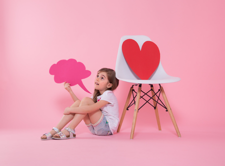 Cute little girl with a speech icon in her hands on a colored background, sitting near a chair with a big red paper heart .The concept of communication . Studio shootingの写真素材