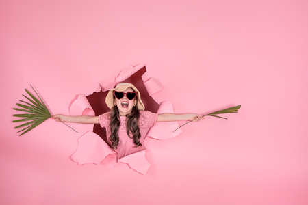 funny little girl looks out from the hole in a beach hat and glasses in the shape of a heart, in the hands of tropical leaves, on a colored background, a place for text, Studio shootingの写真素材