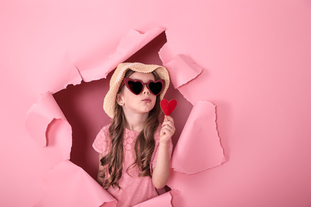 funny little girl looks out from the hole in a beach hat and glasses in the shape of a heart, holding a heart on a stick, on a colored background, a place for text, Studio shootingの写真素材