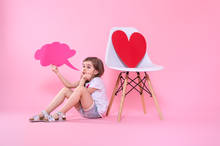 Cute little girl with a speech icon in her hands on a colored background, sitting near a chair with a big red paper heart .The concept of communication . Studio shootingの写真素材