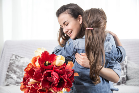 Happy mother's Day. Little cute daughter hugs her mother , and gives a bouquet of tulips, close-up photo. In the interior of the living room, the concept of a happy family lifeの写真素材
