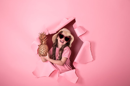 funny little girl peeking out of a hole in a beach hat and heart-shaped glasses with pineapple in her hands, on a colored background, space for text, Studio shootingの写真素材