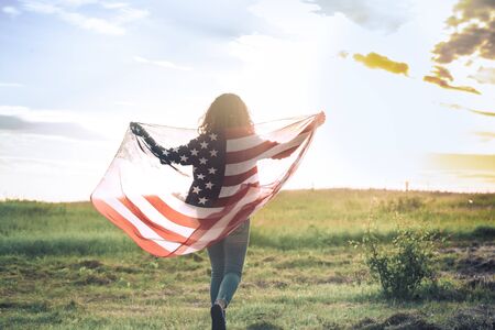Young happy girl running and jumping carefree with open arms over wheat field. Holding USA flag. Toned image. Selective focus.の写真素材