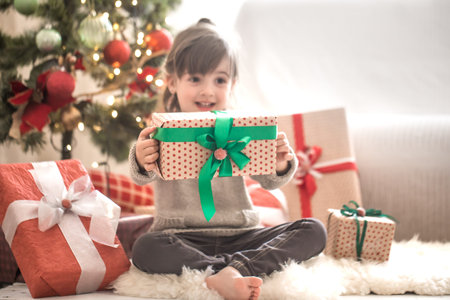 Pretty little girl is holding a gift box and smiling while sitting on her bed in her room at home....