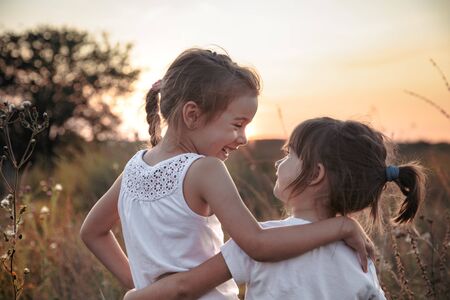 Two little sisters hugging in a field at sunset . Dressed in white. The concept of family values and friendship .の写真素材