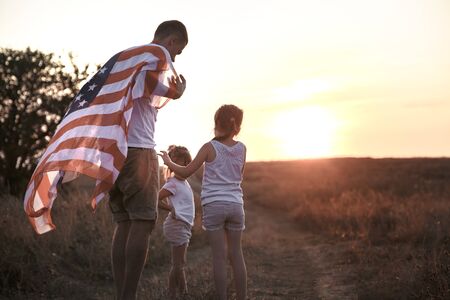 Happy family, dad and daughter holding the American flag at sunset. Dressed in white. The concept of family values and friendship . Patriotic feeling.の写真素材