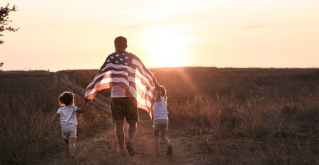 Happy family, dad and daughter holding the American flag at sunset. Dressed in white. The concept of family values and friendship . Patriotic feeling.の写真素材