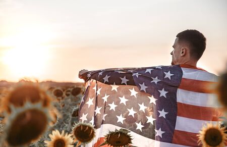 a man holds an American flag at sunset in a beautiful field. Dressed in white. Patriotic feeling.の写真素材