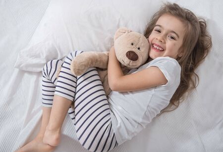 Cute little girl in white bed playing with soft bear toy . Concept of sleep and development of children .の写真素材