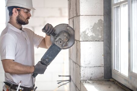 The industrial Builder works with a professional angle grinder to cut bricks and build interior walls. Professional on the construction site. The concept of electrician and handyman.の写真素材