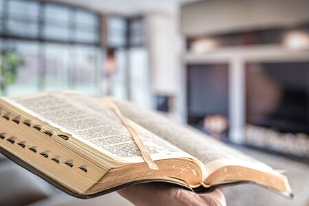 A man holds a Bible against the background of the living room. Reading a book in a cozy atmosphere. Close up.の写真素材