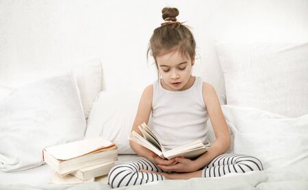 Cute little girl reading a book on the bed in the bedroom. The concept of education and family values .の写真素材
