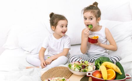 Healthy food at home. Happy two cute children eating fruits and vegetables in the bedroom on the bed. Healthy food for children and teenagers.の写真素材