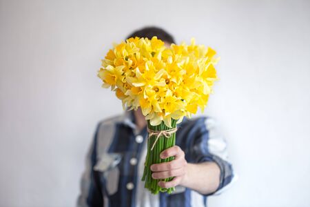 A man in a shirt covers his face with a bouquet of flowers. On light background. The concept of greetings and women's day.の写真素材