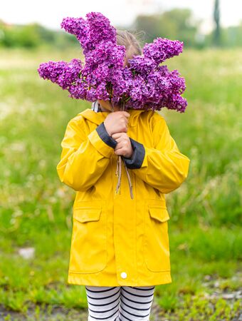A little girl in a yellow jacket covers her face with a bouquet of Lilacs. Creative concept.の写真素材