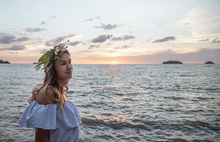 Beautiful young woman with a wreath of flowers on her head by the ocean at sunset.の写真素材