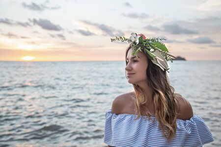 Beautiful young woman with a wreath of flowers on her head by the ocean at sunset.の写真素材