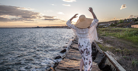 Beautiful young woman by the sea at sunset. Dressed in a long skirt and hat.