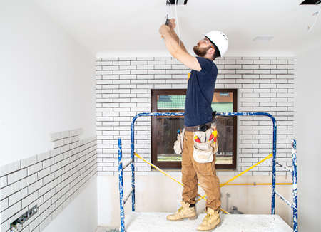 Electrician Builder at work, installation of lamps at height. Professional in overalls with an electrical tool. On the background of the repair site. The concept of working as a professional.の写真素材