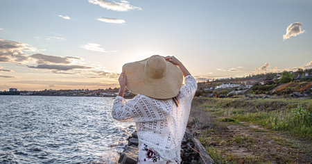 Beautiful young woman by the sea at sunset. Dressed in a long skirt and hat.
