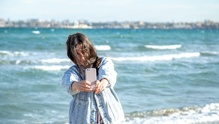 A young stylish woman takes a selfie photo by the sea on the beach.
