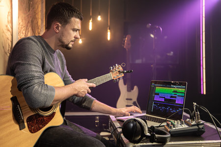 A man with a guitar records sound in a music studio using a special program on his laptop against a...