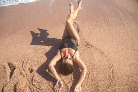 Beautiful woman in a black swimsuit lying on the sand by the sea, top view. The concept of relaxation and vacationの写真素材