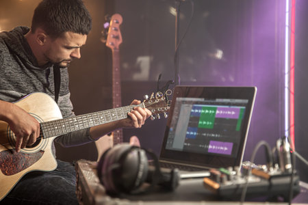 Professional musician recording in studio. Guitarist sitting in front laptop monitor in recording...