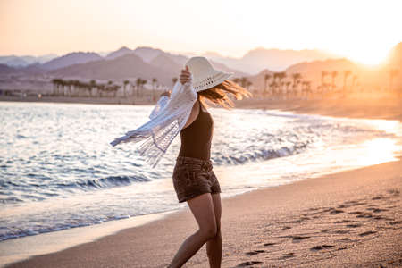 A beautiful boho model in a white Cape and swimsuit poses on the beach in the sunlightの写真素材