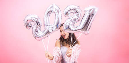Cute young girl with a smile in a festive outfit posing against a pink studio background and holding silver balloons from 2021 numbersの写真素材