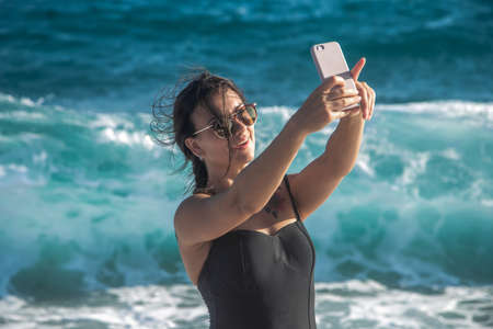A cheerful woman in a swimsuit takes a selfie against the background of sea waves.の写真素材