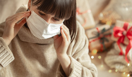Young woman with a mask on her face in a cozy beige sweater on a blurred background with bokeh. The...