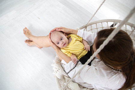 A newborn boy in a yellow T-shirt lies in a hammock chair on the lap of his mother and looks on a...