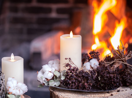 Beautiful candles decorated with dried flowers and cotton are burning near the fireplace. The concept of a cozy holiday.の写真素材