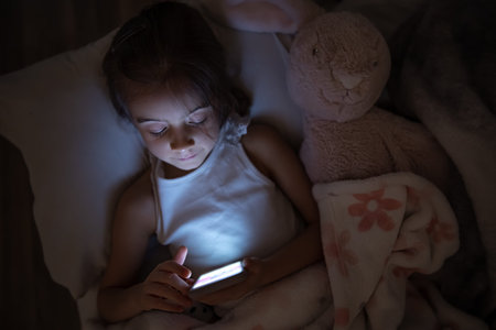 A baby girl lies in bed with a soft toy and uses a smartphone. The concept of child addiction to...