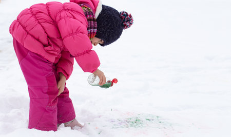 A cute girl with a bottle of paint draws on the snow. The concept of children's creativity and winter fun.の写真素材