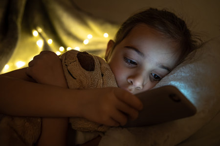 A cute baby girl lies in bed with a teddy bear and uses a smartphone.
