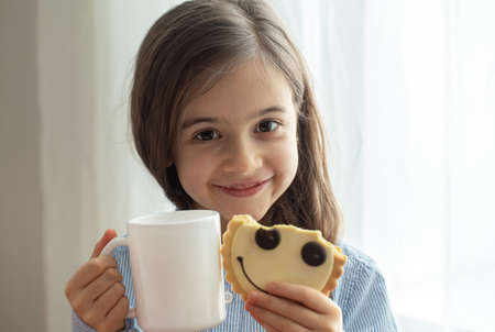 An elementary school girl is having breakfast with milk and funny cookies in the form of a smiley.の写真素材