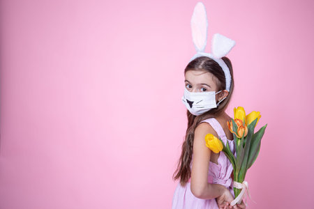 Little girl with easter bunny ears and wearing a medical face mask holds a bouquet of tulips in her...