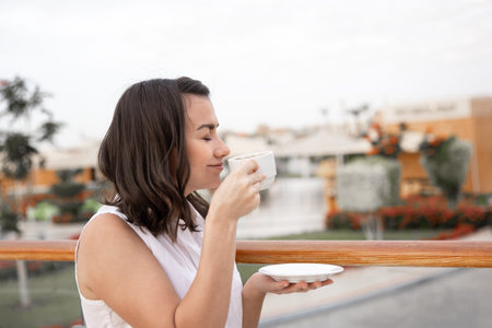 Attractive young woman enjoying the morning outdoors with a cup of coffee and a saucer in her hand....