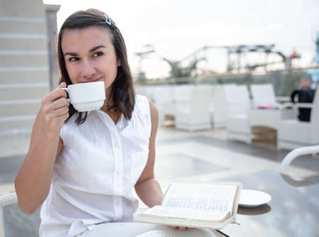 Attractive young woman enjoying morning coffee on an open summer terrace with a book in her hands. Rest and relaxation concept.の写真素材