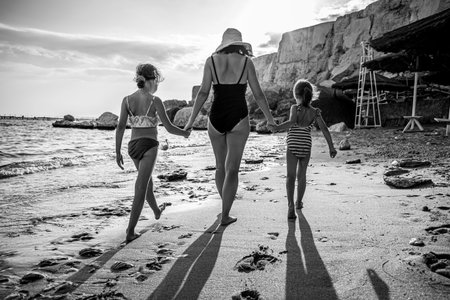 Mom and two little daughters walk along the seashore in swimsuits, holding hands. Vee from the back.