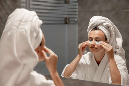 A girl a bathrobe and with a towel on her head sticks patches under her eyes in the bathroom in front of the mirror.の写真素材