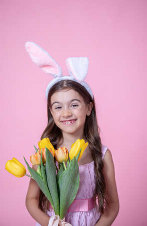 Cheerful little girl with Easter bunny ears smiles and holds a bouquet of tulips in her hands on a pink studio background.の写真素材