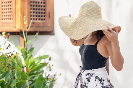 A girl in a large straw hat near the facade of an old house on a hot summer day.