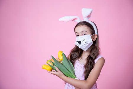 Little girl with easter bunny ears and wearing a medical face mask holds a bouquet of tulips in her hands on a pink studio background.の写真素材